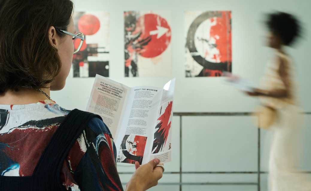 Pessoa de costas a ler um folheto numa galeria de arte, com obras abstractas vermelhas e pretas na parede e alguém desfocado ao fundo.