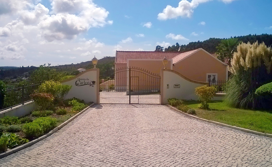Entrada principal da Quinta da Carlota, com portão em ferro, calçada e edifício ao fundo, num ambiente verde e tranquilo.