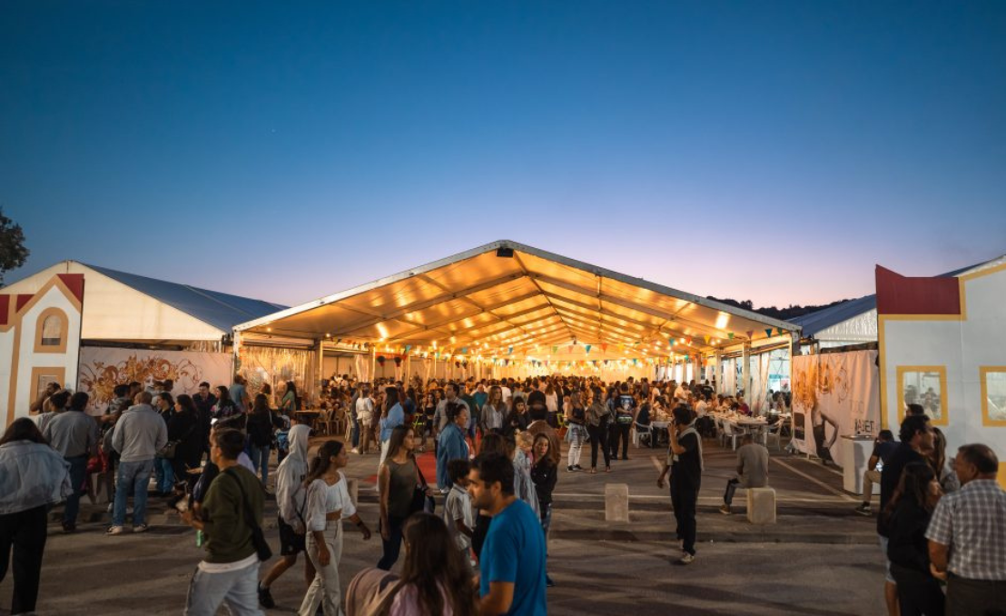 Pessoas a circular numa feira animada com tendas iluminadas ao entardecer